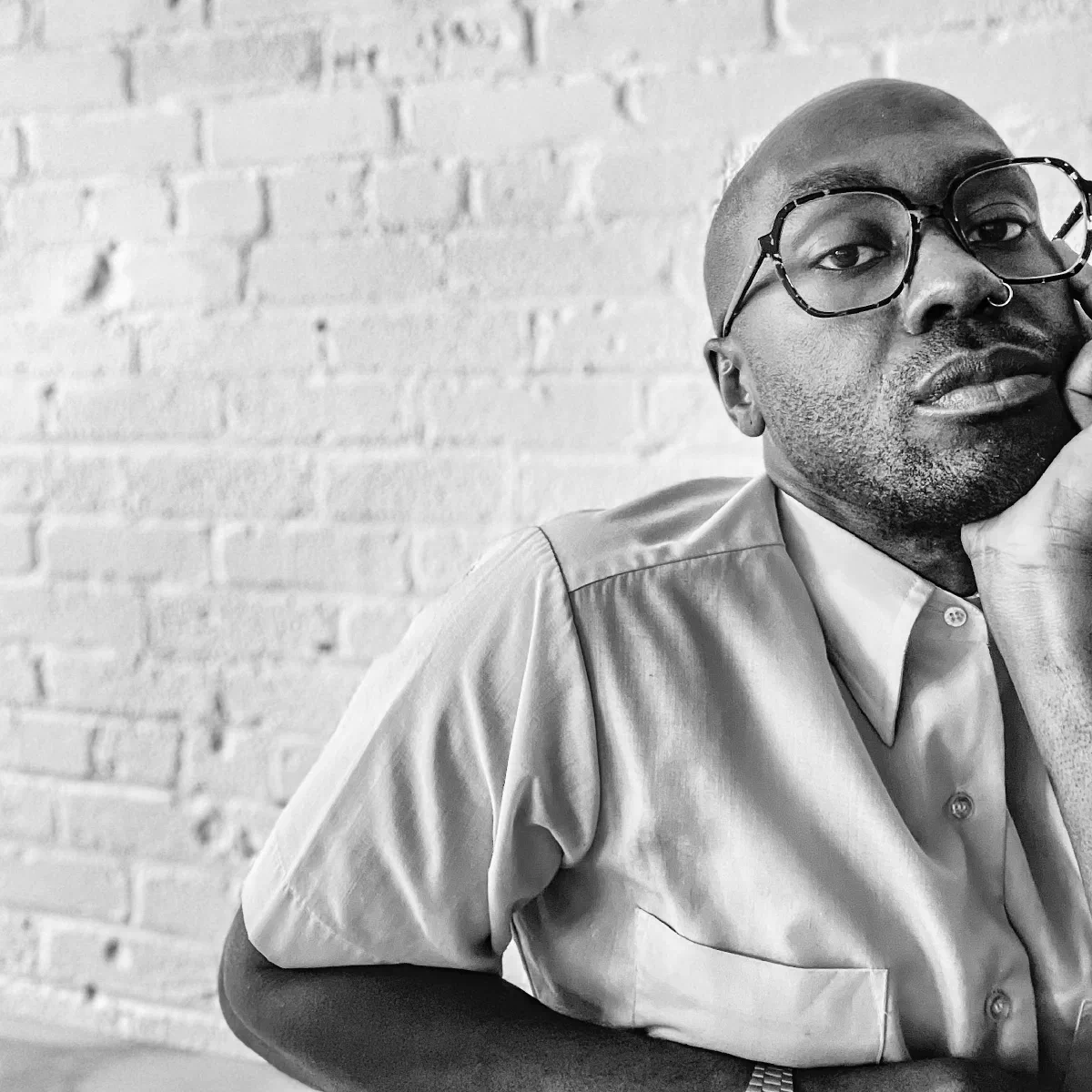Black and white photo of a dark-skinned person in patterned glasses and a light-colored button-up shirt; his head resting on one hand against a painted brick wall.