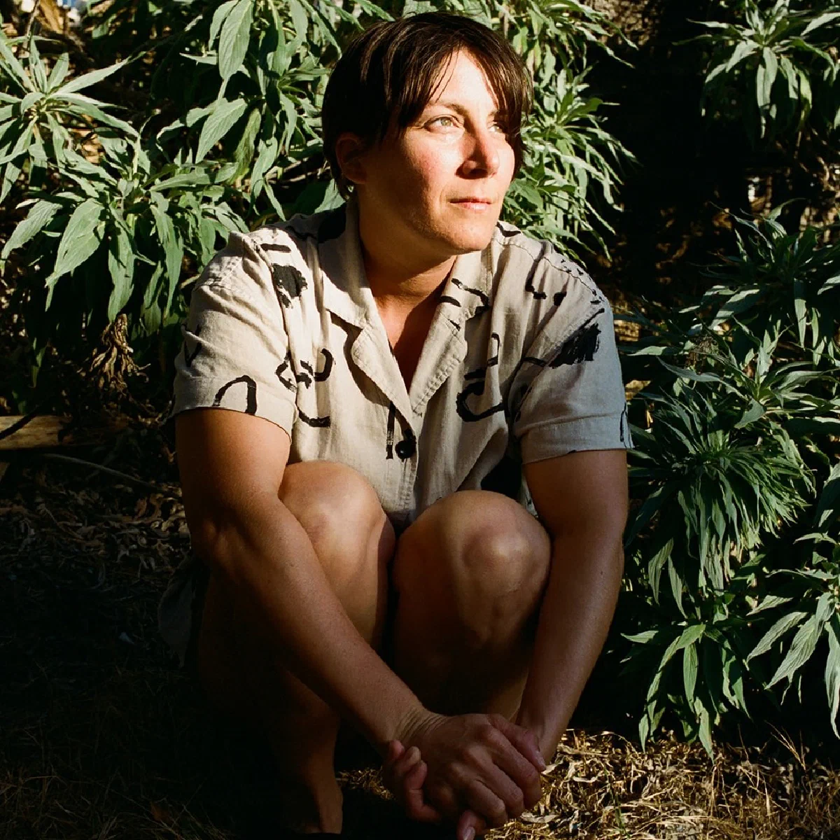 Light-skinned person crouched among green plants in dappled sunlight; she is wearing a taupe-colored shirt with black brushstrokes printed on it.